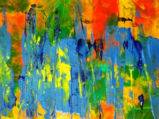 Vibrant abstract painting featuring a mix of blue, yellow, green, and orange colors. Thick brushstrokes create vertical patterns and texture across the canvas. Energetic use of color and impasto technique gives the artwork a dynamic and lively feel.