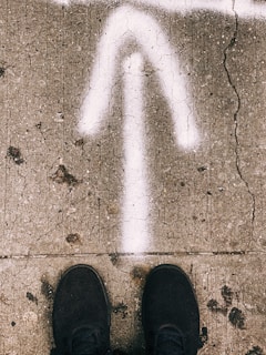 A cracked concrete pavement with a white arrow painted pointing upwards. Black shoes are visible at the bottom, suggesting someone standing on the pavement.