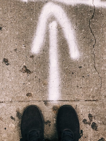 A cracked concrete pavement with a white arrow painted pointing upwards. Black shoes are visible at the bottom, suggesting someone standing on the pavement.