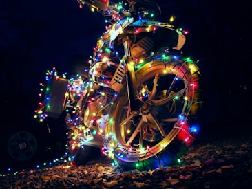 A motorcycle adorned with colorful string lights wrapped around its body and wheels, set against a dark nighttime background with fallen leaves on the ground.