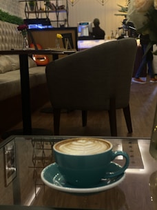 A cozy café scene with a cappuccino in a turquoise cup on a glass table in the foreground. Comfortable seating and wooden flooring complement the warm ambiance. A couple of small vases with flowers are placed on tables, and a person is visible in the background near the counter, creating a relaxed and inviting atmosphere.