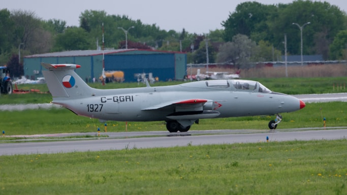 A small jet aircraft is positioned on a runway in an open area, surrounded by a mix of natural greenery and airport facilities. The aircraft has a sleek, gray body with red accents on its tail and wings. A logo or marking is visible on the tail fin. In the background, there are trees and a hangar, indicating an airport environment.