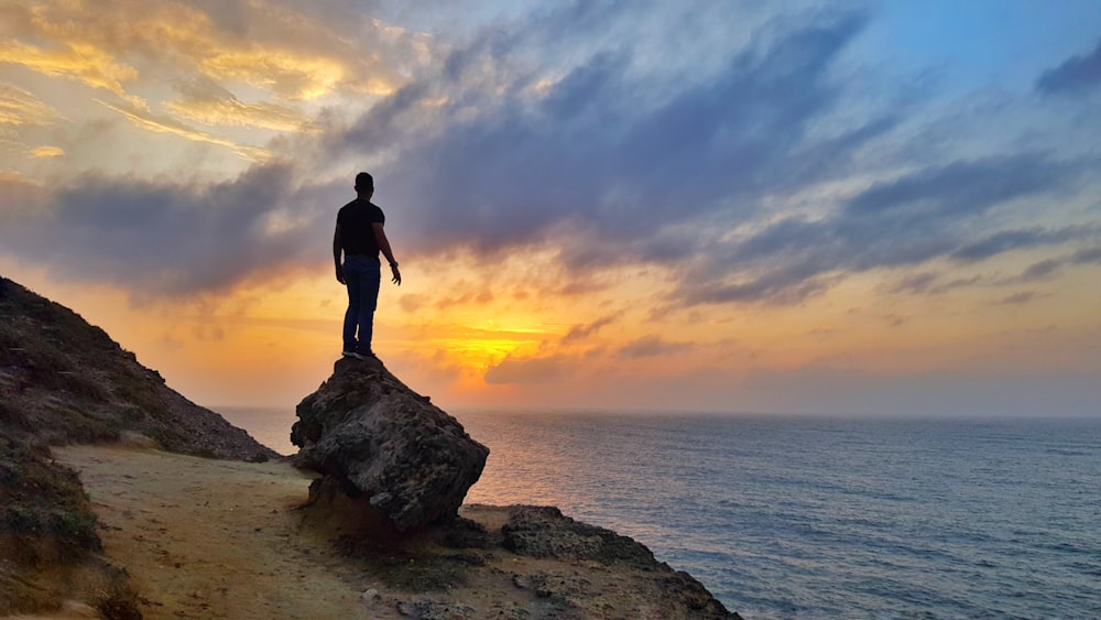 man in black shirt standing on rock near sea during sunset