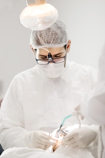 A medical professional wearing protective gear and magnifying glasses is performing a procedure. The setting is brightly lit, highlighting the focus on the detailed work being done. Instruments and gloved hands are visible, indicating a clinical environment.