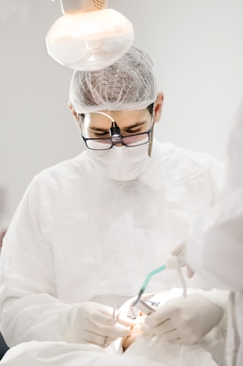 A medical professional wearing protective gear and magnifying glasses is performing a procedure. The setting is brightly lit, highlighting the focus on the detailed work being done. Instruments and gloved hands are visible, indicating a clinical environment.