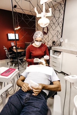 A dentist is performing a procedure on a patient reclining in a dental chair. The dentist is wearing a protective cap, mask, and gloves while using dental tools. The room features modern decor with geometric patterns on the wall, and there is a dental light overhead. Nearby, there is a table with dental instruments and a monitor on the wall.