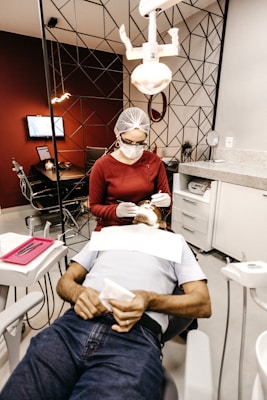 A dentist is performing a procedure on a patient reclining in a dental chair. The dentist is wearing a protective cap, mask, and gloves while using dental tools. The room features modern decor with geometric patterns on the wall, and there is a dental light overhead. Nearby, there is a table with dental instruments and a monitor on the wall.