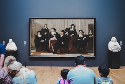 A group of people sits on the floor in front of a large painting displayed in a museum or gallery. The painting depicts several men in historical attire. On either side of the painting, there are marble busts displayed on pedestals.