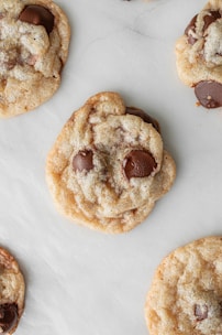 Several chocolate chip cookies are laid out on a white surface, showcasing their golden-brown color and rich chocolate pieces.