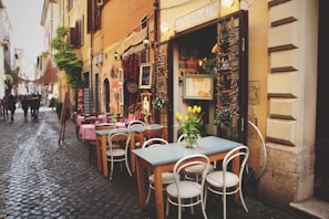 A charming cobblestone street lined with small shops and a cozy outdoor cafe. The tables are set with chairs, some adorned with pink tablecloths. A vase of yellow tulips is placed on one table, adding a touch of color. The storefronts have a rustic appeal with warm, earthy tones and greenery. People are seen walking down the street, contributing to a lively yet relaxed atmosphere.