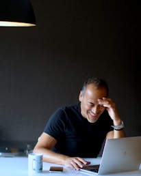 A person is sitting at a table, laughing while looking at a laptop. They have one hand on their forehead and the other on the laptop. There is a coffee mug and a smartphone on the table. A black lamp hangs from above, casting light on the scene.