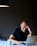 A person is sitting at a table, laughing while looking at a laptop. They have one hand on their forehead and the other on the laptop. There is a coffee mug and a smartphone on the table. A black lamp hangs from above, casting light on the scene.