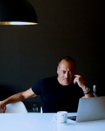 A man sitting at a white table with a laptop, appearing thoughtful. He rests his right elbow on the table, his fingers touching his temple. A white mug with black text is in the foreground. The background features a dark wall and a portion of a black hanging lamp.