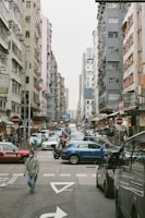 A busy urban street scene with tall residential buildings on both sides. Numerous vehicles, including cars and taxis, are visible on the road, creating a congested traffic situation. People are walking on the street, some wearing face masks. Street signs, shop signs, and various storefronts add to the hustle and bustle of city life.