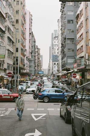 A busy urban street scene with tall residential buildings on both sides. Numerous vehicles, including cars and taxis, are visible on the road, creating a congested traffic situation. People are walking on the street, some wearing face masks. Street signs, shop signs, and various storefronts add to the hustle and bustle of city life.