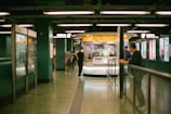 A busy underground station with people walking and signs displayed above. The station is brightly lit with advertisements along the walls and a service center booth in the middle.