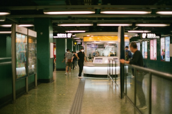 A busy underground station with people walking and signs displayed above. The station is brightly lit with advertisements along the walls and a service center booth in the middle.