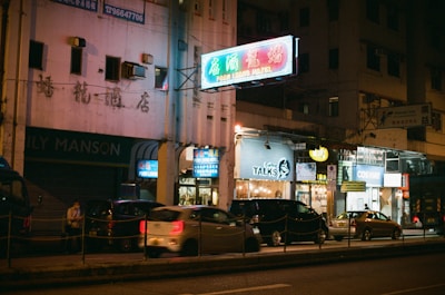 A lively street scene at night featuring various illuminated signs in both English and Chinese. Vehicles are parked along the road, and there are shops and signs including 'Poon Loon Hotel', 'Coffee Talks', and 'Cosway'. The lighting creates a vibrant and bustling atmosphere typical of an urban night setting.