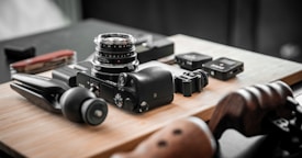 A collection of camera equipment and accessories is arranged on a wooden surface. Prominent items include a camera with a lens attached, a small tripod, and several other small electronic devices. The setting appears organized and focused, highlighting the tools for photography.