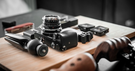 A collection of camera equipment and accessories is arranged on a wooden surface. Prominent items include a camera with a lens attached, a small tripod, and several other small electronic devices. The setting appears organized and focused, highlighting the tools for photography.