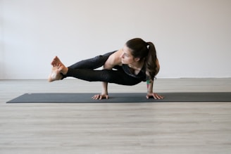 A person is performing a yoga pose, balancing on their hands with legs extended to the side. The environment is minimalistic with a light-colored wooden floor and a plain wall in the background.