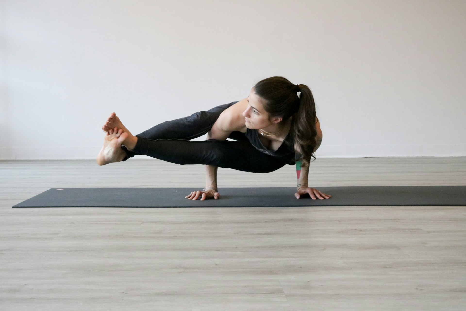 Woman balancing in eight angle pose on a dark mat, both legs extended to one side and crossed at the ankles