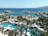 A lively seaside city scene with a vibrant blue ocean visible in the background, dotted with several ships near the horizon. The foreground features a busy resort area with swimming pools surrounded by palm trees and numerous lounging areas with white sunshades. Many people are enjoying the pools while the marina in the midground is filled with boats docked at the piers.