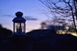 A lantern with a lit candle inside rests on a surface. It is silhouetted against a twilight sky. The background shows dark silhouettes of trees and structures, with the sky transitioning from blue to soft purple and orange hues at the horizon.