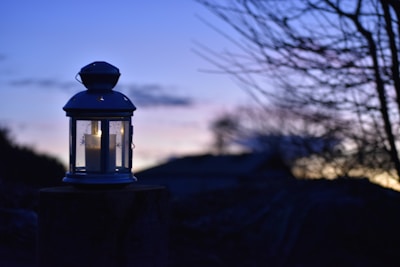 A lantern with a lit candle inside rests on a surface. It is silhouetted against a twilight sky. The background shows dark silhouettes of trees and structures, with the sky transitioning from blue to soft purple and orange hues at the horizon.