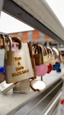 An assortment of padlocks attached to a metal railing, some appear rusted while others are distinct with shapes like hearts. One notable padlock displays engraved names and a date.