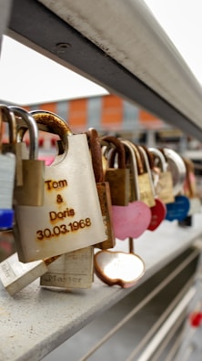 An assortment of padlocks attached to a metal railing, some appear rusted while others are distinct with shapes like hearts. One notable padlock displays engraved names and a date.