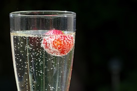 A champagne flute filled with a fizzy, clear liquid, inside which a single ripe strawberry is suspended. The bubbles cling to the surface of the strawberry and rise through the beverage. The glass is set against a dark, out-of-focus background, highlighting the sparkle of the bubbles.