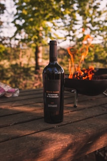 A bottle of Monte Sasso Primitivo di Manduria wine is placed on a wooden table outdoors. Behind the bottle, there is a barbecue grill with bright orange flames flickering upwards. The background is lush with green trees, suggesting a forested or garden setting.