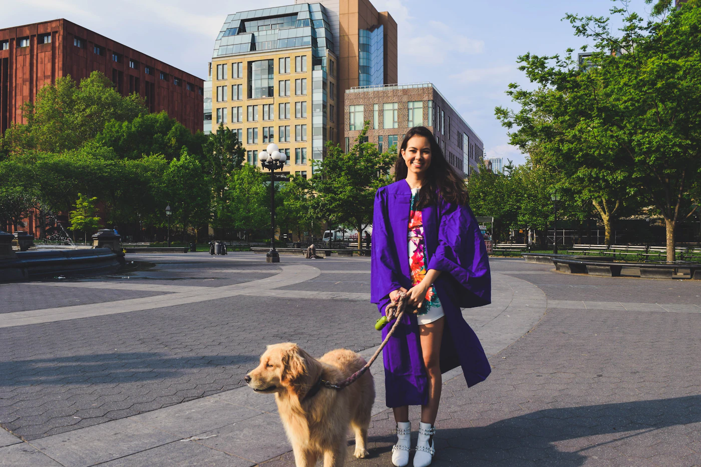 A young graduate in a cap and gown walking a leashed dog across a campus plaza with university buildings behind her