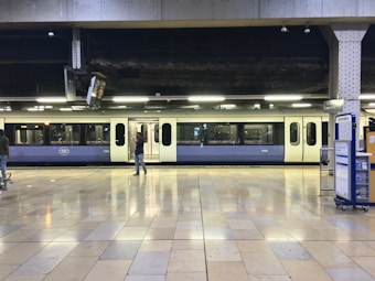 A train is stationed at an indoor platform with people walking nearby. The scene includes a tiled floor, overhead lighting, and signage indicating platform numbers. The train has a light blue and cream color scheme.