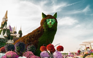 A large topiary sculpture in the shape of a cat is surrounded by vibrant flower arrangements. The cat features foliage forming its fur and green eyes. In the background, a whimsical, fairytale-like castle with cone-topped towers adds to the fantastical atmosphere. The sky is partly cloudy, creating a bright and vivid backdrop.