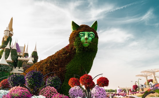 A large topiary sculpture in the shape of a cat is surrounded by vibrant flower arrangements. The cat features foliage forming its fur and green eyes. In the background, a whimsical, fairytale-like castle with cone-topped towers adds to the fantastical atmosphere. The sky is partly cloudy, creating a bright and vivid backdrop.