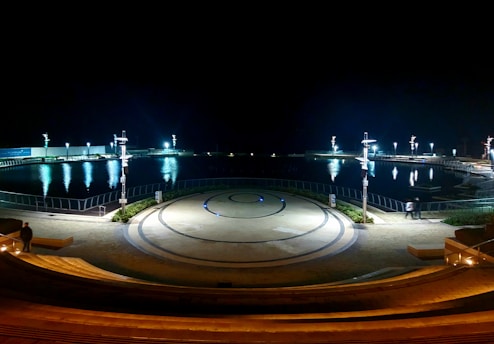 A nighttime scene of a large, circular amphitheater overlooking a body of water. The space is lit by modern streetlamps, casting reflections across the calm water. The steps lead down to a central circular area, marked by concentric patterns on the ground. Sparse greenery is visible near the railings, and a few people appear to be walking in the area.