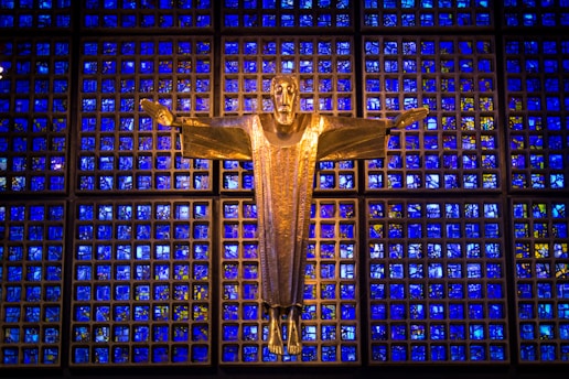 A golden statue of Jesus with outstretched arms is prominently displayed against a striking backdrop of blue stained glass panels, creating a serene and majestic atmosphere.
