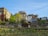 A hillside urban vineyard with several buildings rising in the background. The vineyard rows are neatly arranged with stakes, and there is a variety of lush green vegetation and flowering plants. The buildings have a mix of architectural styles, featuring stone, wood, and glass elements. The sky above is a clear blue.
