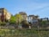 A hillside urban vineyard with several buildings rising in the background. The vineyard rows are neatly arranged with stakes, and there is a variety of lush green vegetation and flowering plants. The buildings have a mix of architectural styles, featuring stone, wood, and glass elements. The sky above is a clear blue.
