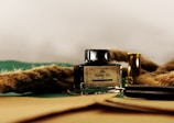 A glass ink bottle labeled 'The Feather Pen' is placed on a surface alongside a thick rope and a small brass container. A black pen or pencil lies nearby, partially visible among the objects. The background is a simple light color, contrasting with the dark and earthy tones of the objects in the foreground.
