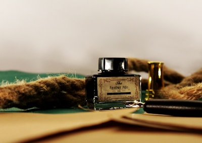 A glass ink bottle labeled 'The Feather Pen' is placed on a surface alongside a thick rope and a small brass container. A black pen or pencil lies nearby, partially visible among the objects. The background is a simple light color, contrasting with the dark and earthy tones of the objects in the foreground.