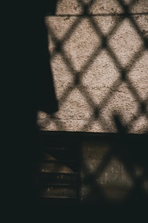 A shadowed lattice pattern is projected onto a textured wall, creating a criss-cross design. The lighting is dim, highlighting the rustic surface quality of both the wall and the partial view of the object behind the shadow.