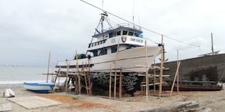 A large fishing boat is on dry land, propped up on wooden supports for maintenance or repairs. The boat is named San Luis III. In the background, a cloudy sky and the ocean are visible, with several small boats on the horizon. The sand is scattered with various items related to boat maintenance.
