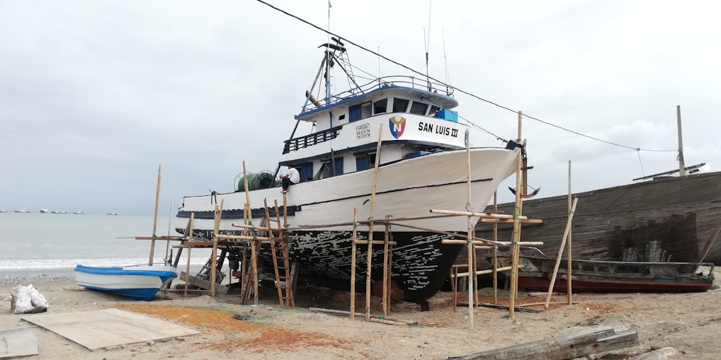 A large fishing boat is on dry land, propped up on wooden supports for maintenance or repairs. The boat is named San Luis III. In the background, a cloudy sky and the ocean are visible, with several small boats on the horizon. The sand is scattered with various items related to boat maintenance.