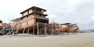 Several large wooden ships are under construction on a sandy beach. The structure is supported by scaffolding and surrounded by construction materials. The setting appears to be near a coastal area with cloudy skies overhead.