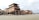 Several large wooden ships are under construction on a sandy beach. The structure is supported by scaffolding and surrounded by construction materials. The setting appears to be near a coastal area with cloudy skies overhead.