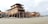 Several large wooden ships are under construction on a sandy beach. The structure is supported by scaffolding and surrounded by construction materials. The setting appears to be near a coastal area with cloudy skies overhead.