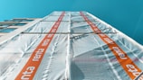 A tall building covered in scaffolding with protective sheeting. The sheeting has a repeating orange stripe with the word 'renta' on it. The building is set against a clear blue sky.